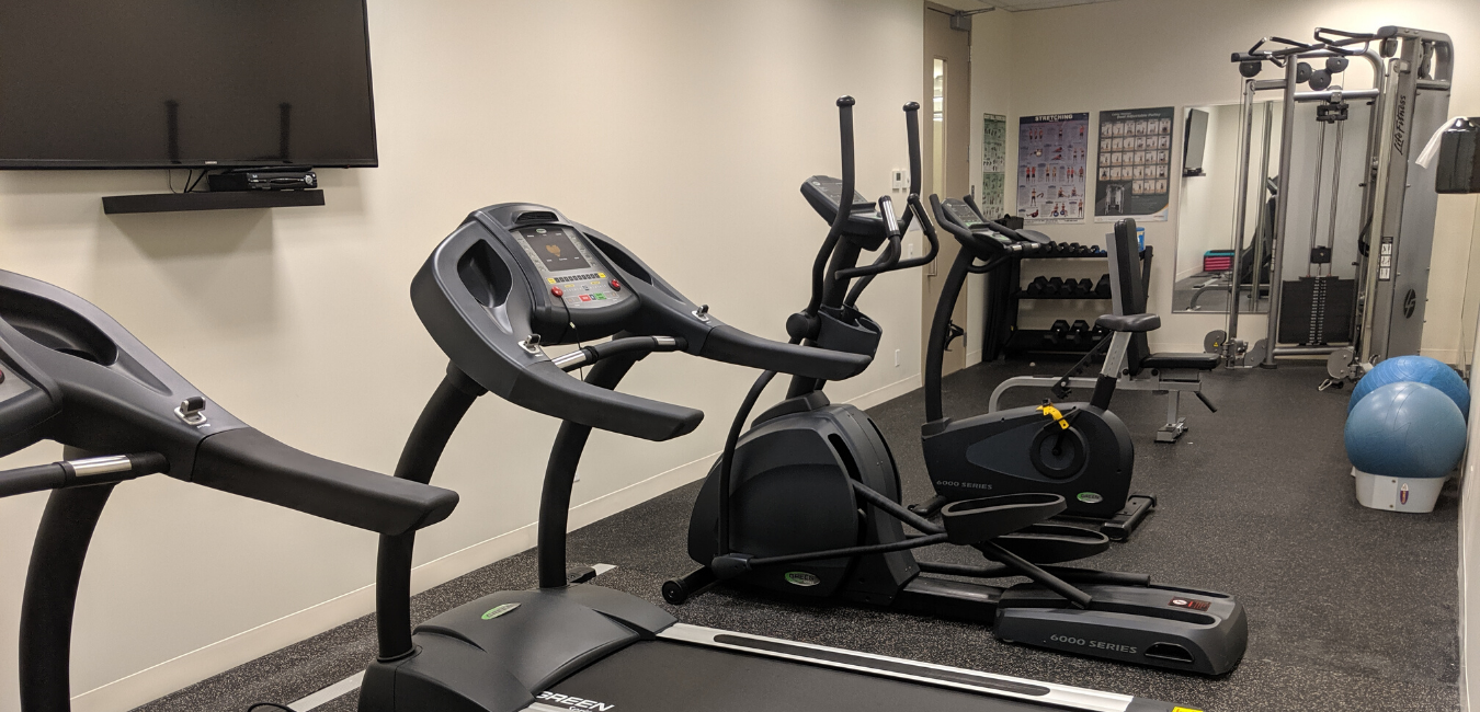 Bennington's one-site fitness room is shown. There's a wall-mounted TV, two treadmills, an elliptical, a stationary bike, free weights, a LifeFitness weight machine and a couple of exercise balls.