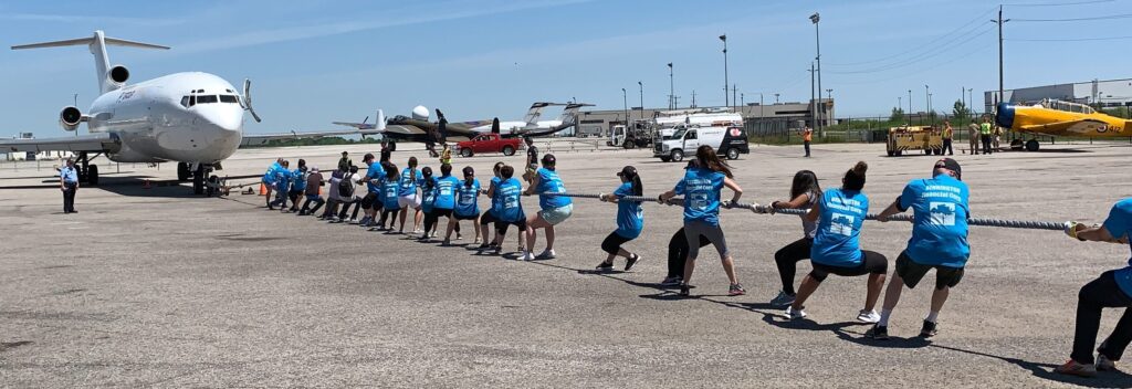 This is a photo of some Bennington team members all pulling on a large rope in unison. The large rope is attached to a plane. They are on the concrete of a tarmac with a white plane in the background. The sky is blue.