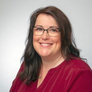 Amanda has a welcoming smile. She is wearing a long sleeve blouse in a raspberry colour and thin black glasses.