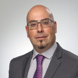 Sergio is wearing a dark gray jacket with a light shirt and a red tie with purple details. He is sporting metallic glasses, a goatee and a slight smile.